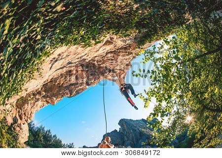 Athletic Man Climbs A Rock, 7c Category Route In The Forest With Rope, Against Sunset And The Other 