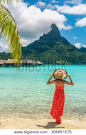 Bora Bora Tahiti exotic tropical vacation destination tourist woman on ocean beach at view of Mt Otemanu in French Polynesia. Girl in red dress and hat relax on honeymoon getaway.