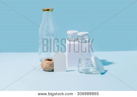 Composition Of Glass Jars On Pedestal, Empty Milk Bottle, Hank Of Twine And Blank Tag Over Blue Back
