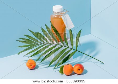 Peach Juice In A Bottle On White Paper Pedestal And Fresh Apricots At Foundation And Palm Branch Lea