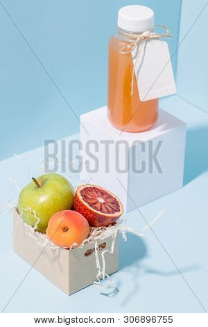 Peach Juice In A Bottle On Pedestal And Fresh Apricot, Apple And Blood Orange Resting In Disposable,