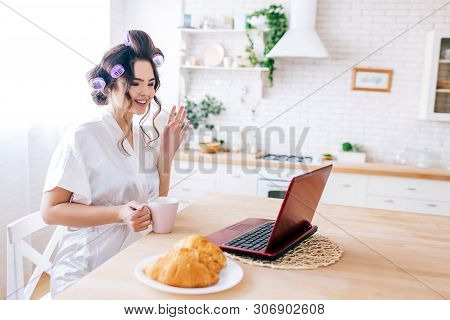 Young Carefree Housekeeper Sitting At Table In Kitchen. Wave Hand During Video Chat. Holding White C