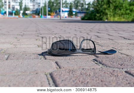 Broken Sun Glasses Lie On A Stone Tiled Walkway In The City Park Against The Background Of Residenti
