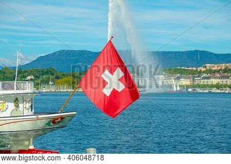 Swiss Flag In Foreground And Jet Deau Fountain In Geneva Lake On Background, The Most Famous Attract