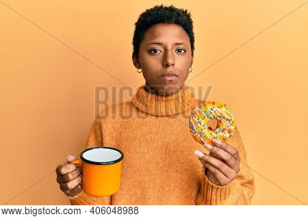 Young african american girl eating doughnut and drinking coffee relaxed with serious expression on face. simple and natural looking at the camera. 