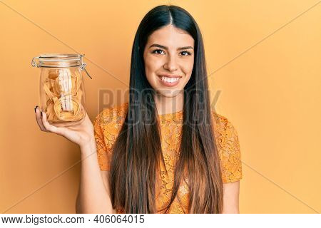 Young hispanic woman holding jar with uncooked pasta looking positive and happy standing and smiling with a confident smile showing teeth 