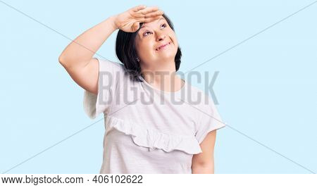 Brunette woman with down syndrome wearing casual white tshirt very happy and smiling looking far away with hand over head. searching concept. 