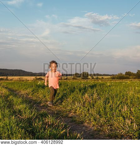 Little Curly Boy In Orange T-shirt Running In A Meadow, Sunset Light. Beautiful Landscape In Summer,