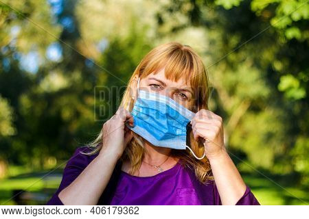 Front View Close-up Of Middle-aged Caucasian Blonde Woman Putting Blue Medical Mask Standing In A Gr