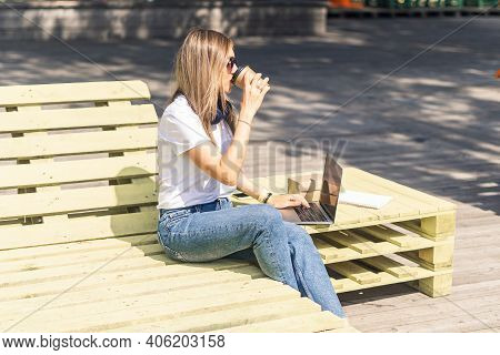 Beautiful Woman Working At Notebook Sit Down On Bench Outside And Drinking Coffee. Using Computer An