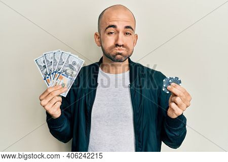 Young hispanic man playing poker holding dollars banknotes and chips puffing cheeks with funny face. mouth inflated with air, catching air. 