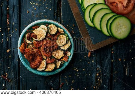 high angle view of a rustic green plate with some slices of dried tomato and zucchini, served as appetizer or snack, and some slices of raw tomato and zucchini, on a dark gray rustic wooden surface