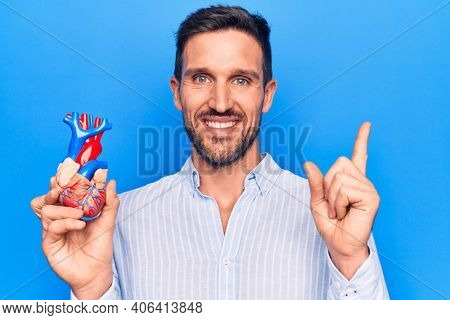 Young handsome man holding heart organ with veins and arteries over blule background smiling happy pointing with hand and finger to the side
