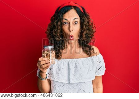 Middle age hispanic woman holding jar with walnuts scared and amazed with open mouth for surprise, disbelief face 