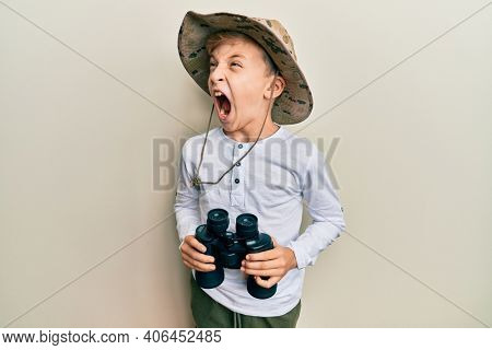 Little caucasian boy kid wearing explorer hat holding binoculars angry and mad screaming frustrated and furious, shouting with anger. rage and aggressive concept. 