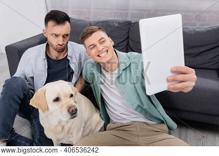 Happy Same Sex Couple Taking Selfie On Digital Tablet Near Labrador In Living Room.
