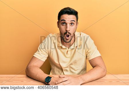 Young hispanic man wearing casual clothes sitting on the table afraid and shocked with surprise expression, fear and excited face. 