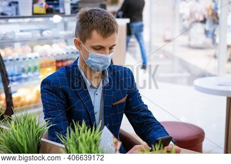 A Young Man In A Jacket Sits In A Cafe In A Medical Mask With A Phone In His Hand