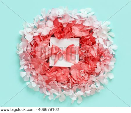White Gift Box With A Bow Between Pink Flowers Top View On Light Green Background
