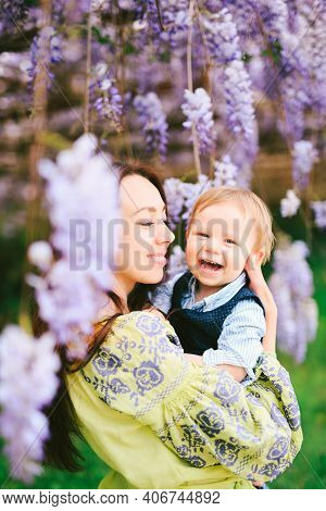 Cute Red-haired Baby Boy Laughing In His Mothers Arms Under A Wysteria Tree