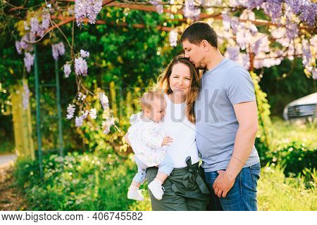 Man And Woman Holding Their Baby Girl Under A Wysteria Tree In The Park
