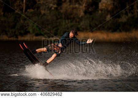 Handsome Sporty Man In Wetsuit Holds Taut Rope And Rides Wakeboard On The Water Surface