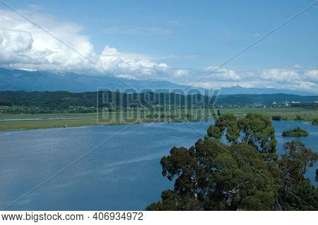 An Ancient Relict Lake In Pitsunda. Mountains Covered With Snow In The Background. Huge White Clouds