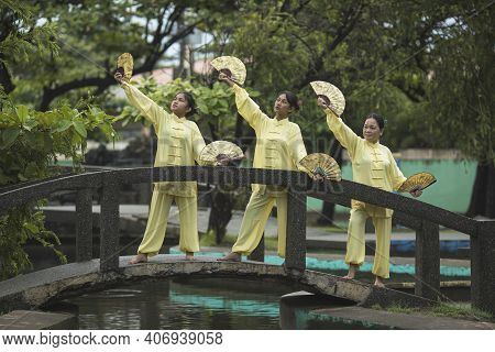 Group Of Beautiful Young Asian Women Practicing Tai Chi With Fans On The Bridge In The Park Wearing 