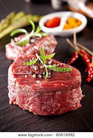 Pieces of red raw meat steaks with rosemary served on black stone surface.