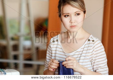 Young Woman Sewing At Home, Hemming Blue Fabric, Stands And Looks Pensively To The Side. Copy Space.