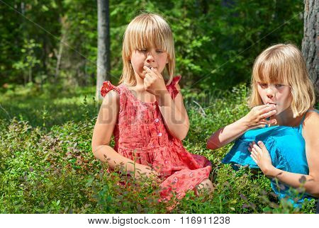 Two little girls wearing blue and red summer dress picking and eating whortleberries in a forest
