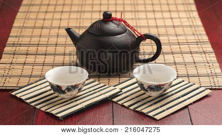 Chinese brown ceramic teapot on bamboo mat. Two white tea cups on a little wooden color mat. Two empty tea porcelans cup. Red table background