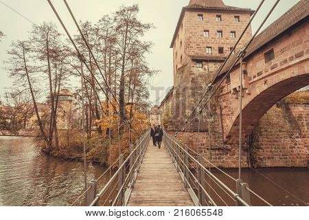 People walk along a wooden suspension bridge across the river in the historic city of Nuremberg with historical walls. Old Bavaria of Germany.