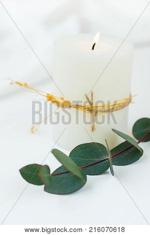 Thick White Lit Candle with Twine and Dry Plant Twig Silver Dollar Eucalyptus White Cloth Background Copy Space. Banner Poster Postcard Template Inspirational
