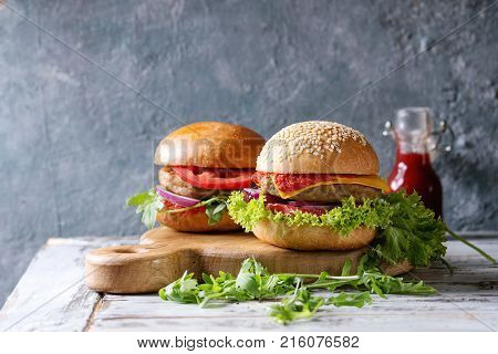 Set of homemade burgers in classic buns with tomato sauce, lettuce, arugula, cheese, onion on wood serving board over white wooden plank table. Rustic style. Homemade fast food.