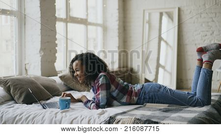 Curly mixed race student girl lauging while using laptop for sharing social media lying in bed at home