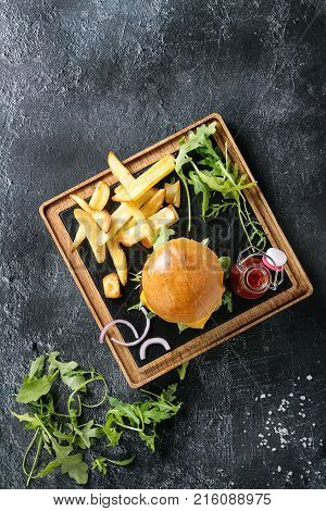 Homemade traditional hamburger with beef, tomato, cheese, aragula, served on wooden slate serving board with french fries and ketchup sauce on dark texture background. Top view with space