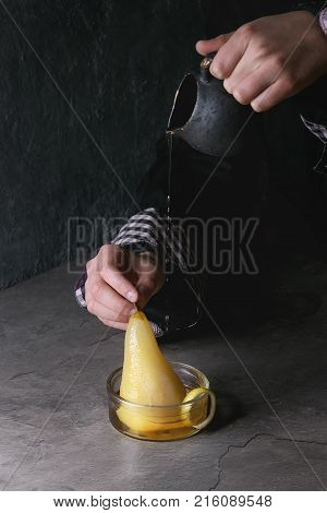 Child hands hold jug and pouring by syrup traditional dessert poached pears in white wine in glass bowls with lemon zest over gray texture table.