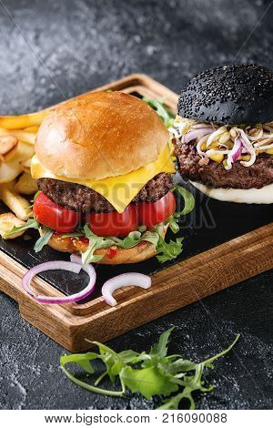Homemade traditional hamburger with beef, tomato, cheese, aragula, served on wooden slate serving board with french fries and ketchup sauce on dark texture background. Close up
