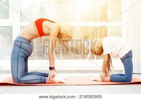 Mother and daughter are engaged in yoga in sportswear. They are in a bright room with panoramic windows. The in a ushtrasana asana, camel pose