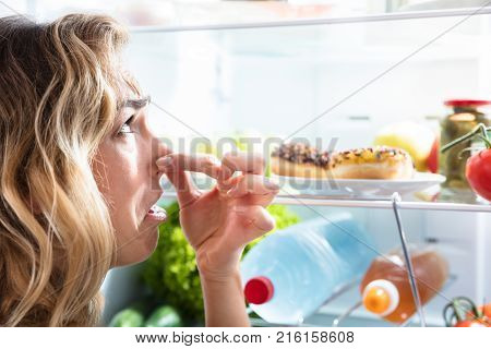 Close-up Of A Young Woman Holding Her Nose Near Foul Food In Refrigerator