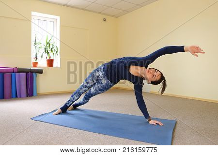 Sportswoman focused on stretch before Pilates class. Girl doing warm up for legs on mat. Female has hair pulled back, concentrated look. Athlete dressed in dark top, yoga legging trains in fitness room. Concept of online training, customizable workouts.