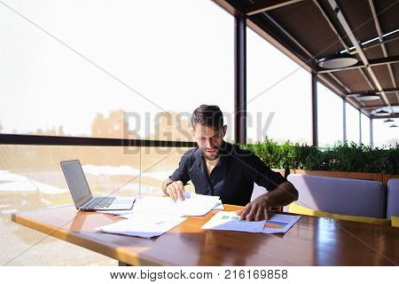 Placeman sorting papers on desk near laptop. Strong male hands with watch near window. Concept of stopping document clutter on workplace.