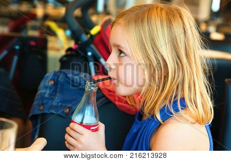 A cute six year old blonde girl in a summer blue dress in the restaurant drinking a soft drink from a bottle with a straw and watches the program on TV turning away from the table