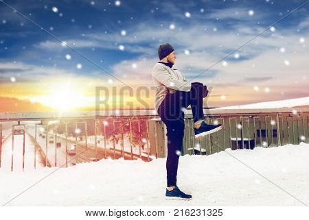 fitness, sport, people, exercising and healthy lifestyle concept - young man stretching leg and warmig up on snow covered winter bridge