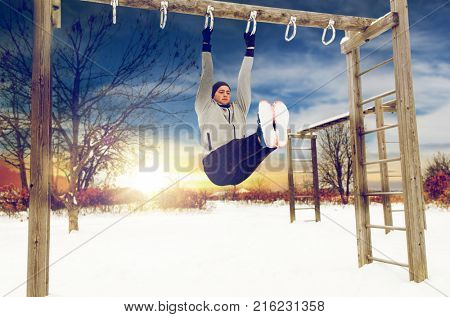 fitness, sport, exercising, training and people concept - young man doing leg pull ups on horizontal bar and flexing abdominal muscles outdoors in winter