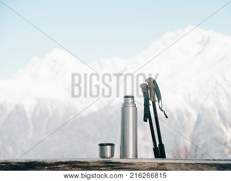 Thermos and trekking poles on wooden table on background of snowy mountains in winter.