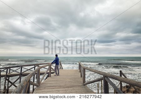 Man On Boardwalk Looking At The Sea In A Clouded Day. Guardamar Beach. Alicante Spain