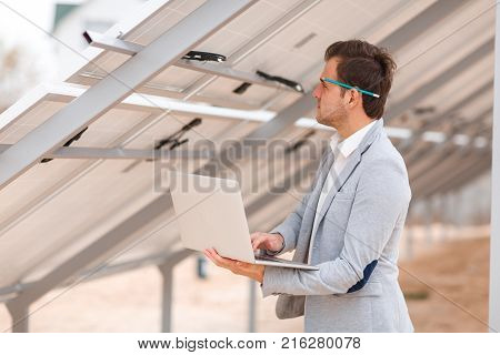 A solid man in a suit with a pencil behind his ear, holds a modern laptop in his hands and looks at the wiring harness for the solar panels installed on the roof. Outdoors