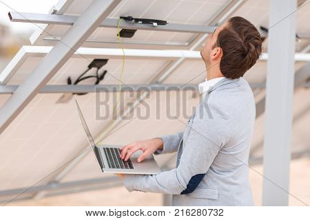 An adult man in a jacket checks the work of solar panels by connecting a modern laptop to a solar cell. Outdoors